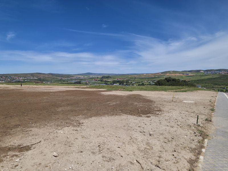 A vacant residential plot featuring a level building site, adjacent to a paved road and sidewalk, offering expansive views of the surrounding landscape.