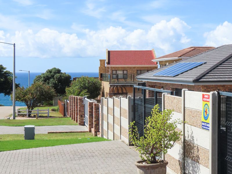 Front exterior view featuring a paved driveway, secure boundary walling, and solar panels installed on the tiled roof with a glimpse of the ocean.