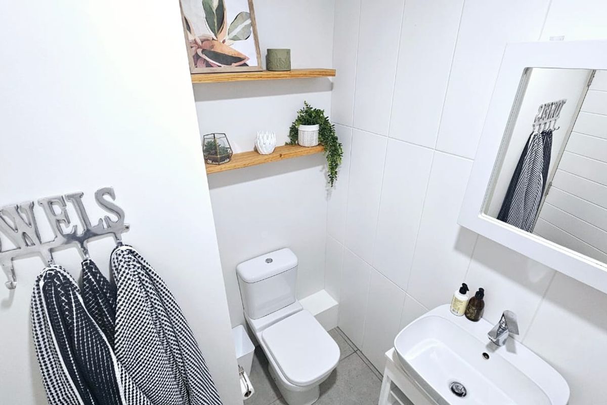 A clean and modern bathroom featuring white tiled walls, a wall-mounted basin with mixer tap, a toilet, and floating timber shelving.