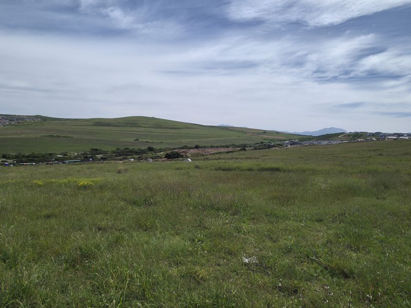 A wide-angle view of an expansive, undeveloped residential plot featuring natural grassland with surrounding hills and distant neighborhood scenery.