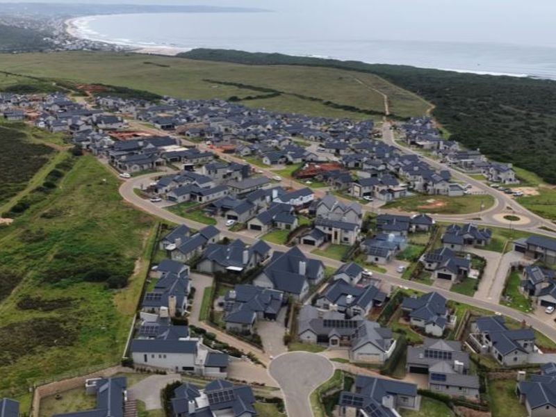 An aerial view of a residential estate situated along the coastline, showcasing the layout of homes, road infrastructure, and surrounding natural landscape.