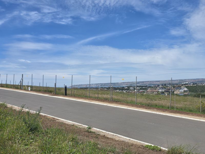A view of a paved residential road with electric perimeter fencing, overlooking a coastal landscape and distant town.