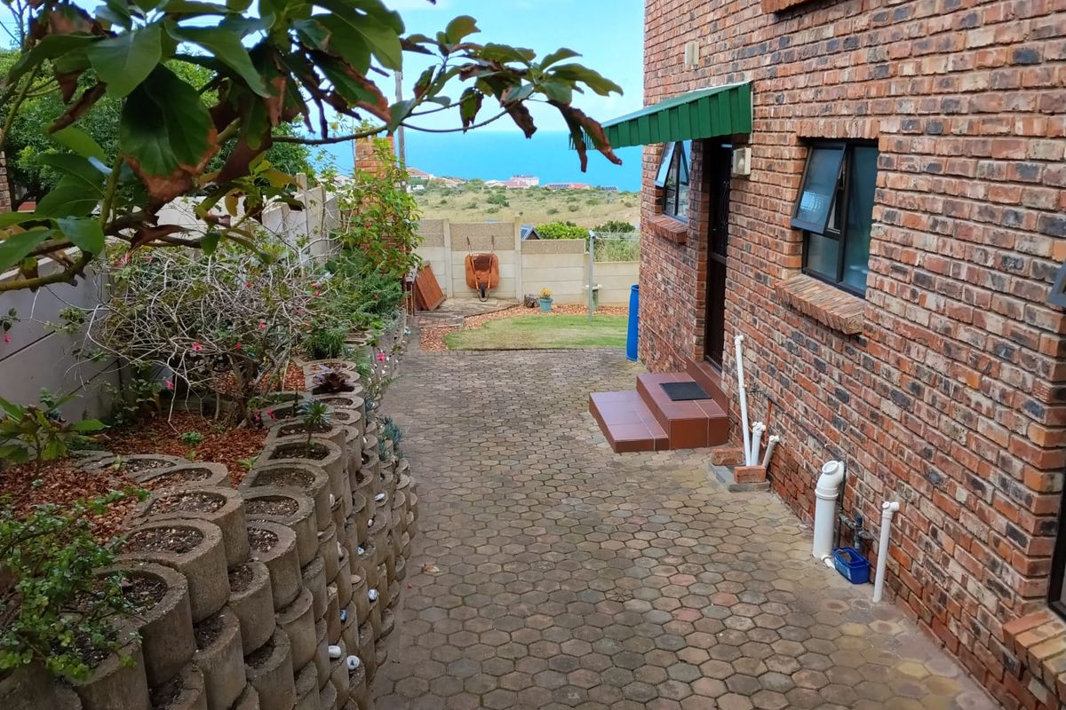 A side exterior view of the house featuring brick facade, paved walkway, tiered garden retaining wall, and a distant ocean view.