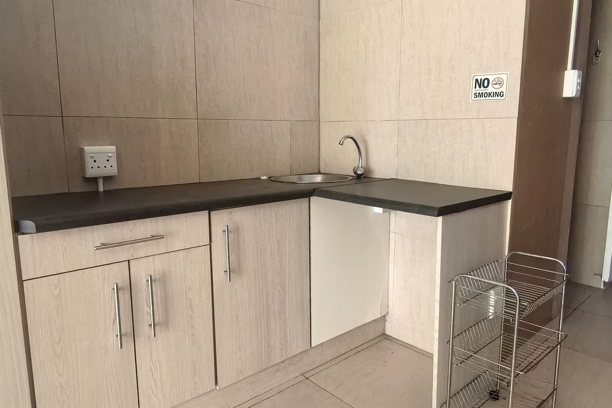 A compact kitchenette area featuring light wood-grain cabinetry, a dark countertop, and a stainless steel sink with tiled splashbacks.