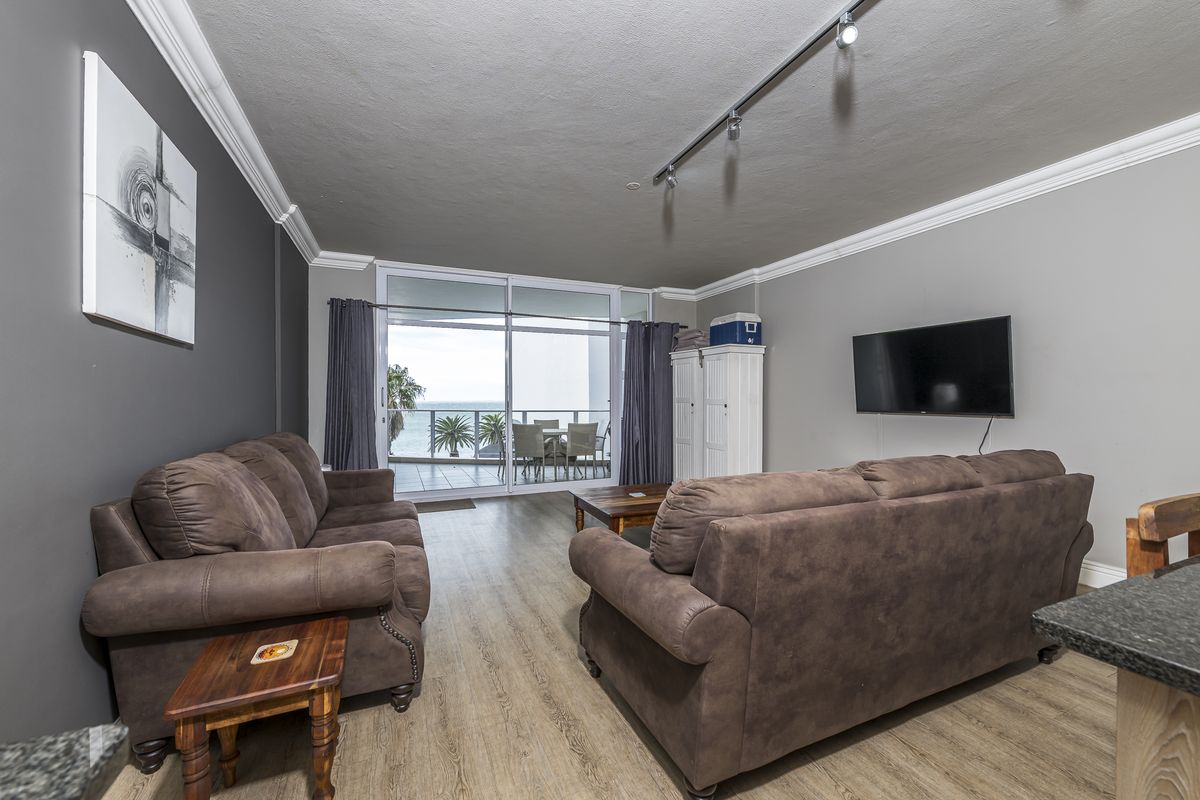 Spacious living room featuring laminate flooring, crown molding, track lighting, and large sliding glass doors leading to a balcony with ocean views.
