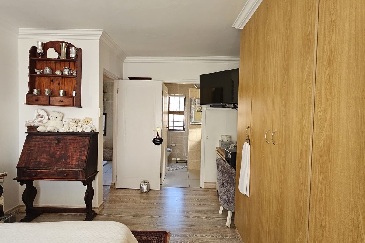 A bedroom featuring light wood-look laminate flooring, extensive built-in cupboards, and coving, with an adjoining en-suite bathroom visible through the doorway.