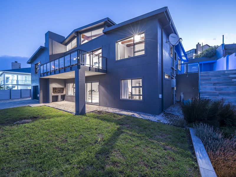 Rear view of a modern multi-story house featuring a spacious balcony, paved patio area with built-in braai, and landscaped garden.