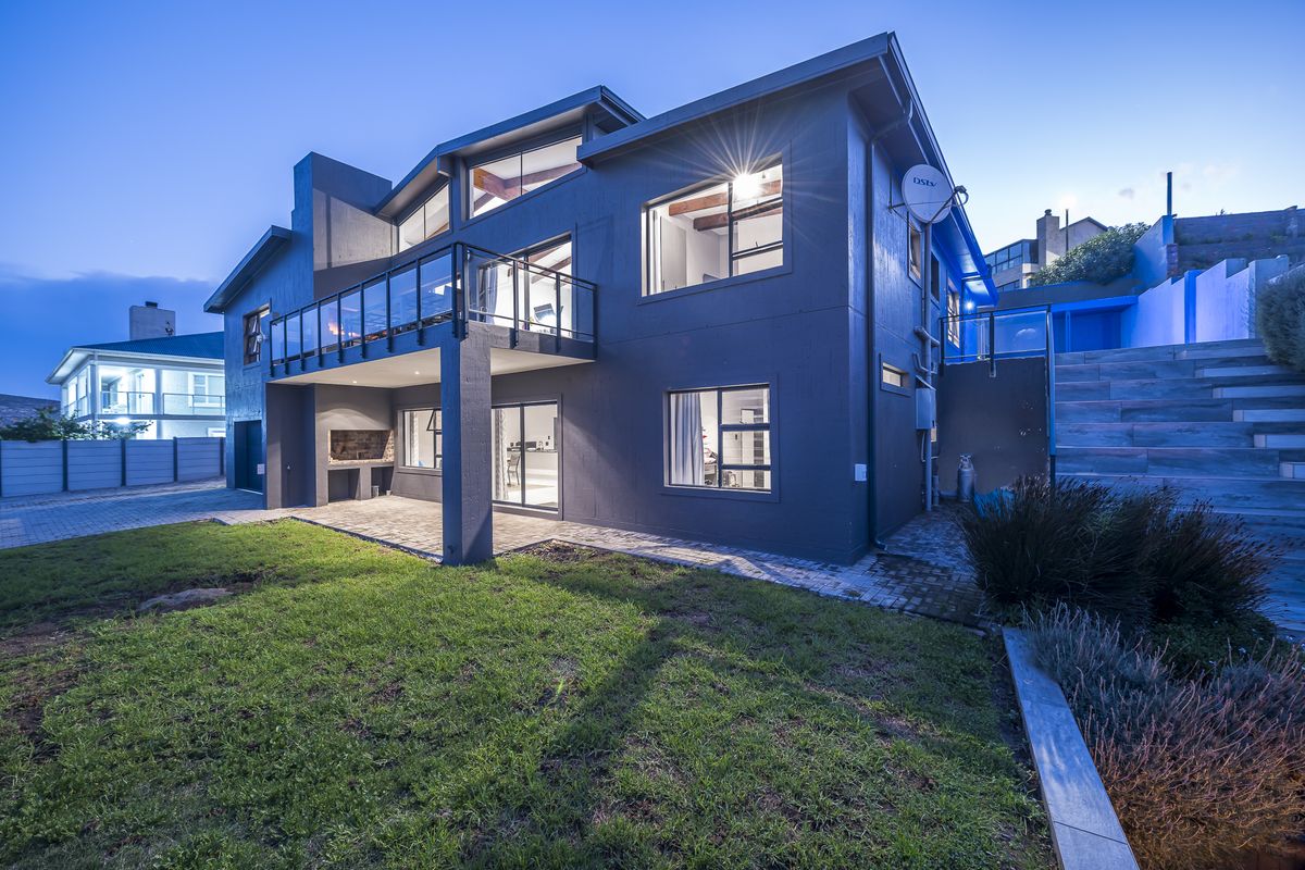 Rear view of a modern multi-story house featuring a spacious balcony, paved patio area with built-in braai, and landscaped garden.