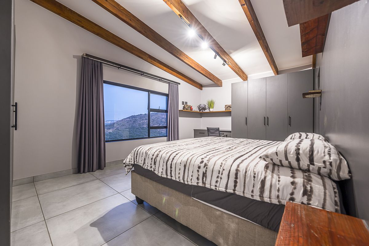 Modern bedroom featuring exposed timber ceiling beams, built-in cabinetry, a dedicated workspace, and large windows offering scenic mountain views.