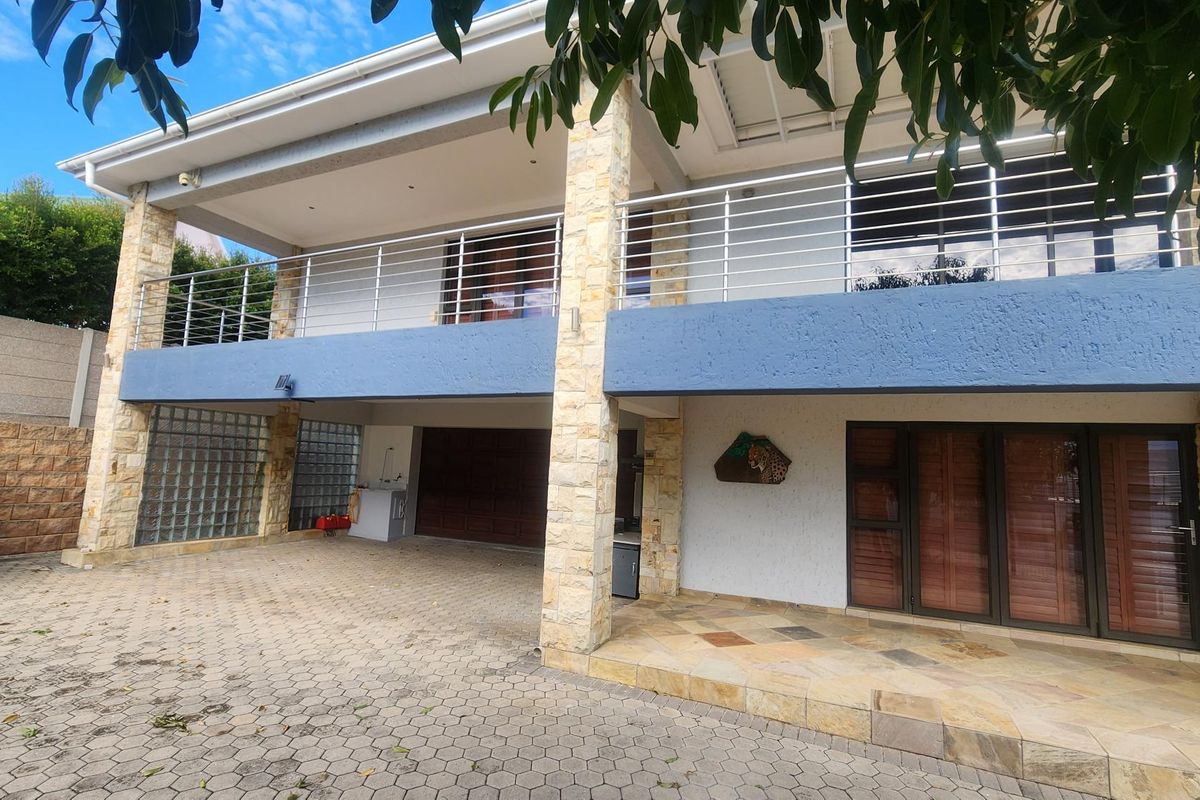 A multi-level exterior view featuring a paved driveway, covered patio area, stone-clad pillars, and a balcony with stainless steel railings.