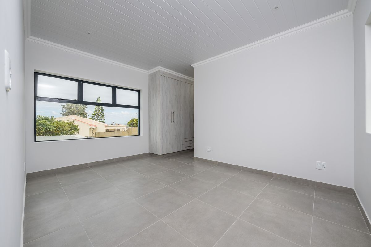 A bright, empty bedroom featuring modern grey floor tiles, built-in cupboards, and a large window with views of the surrounding neighbourhood.