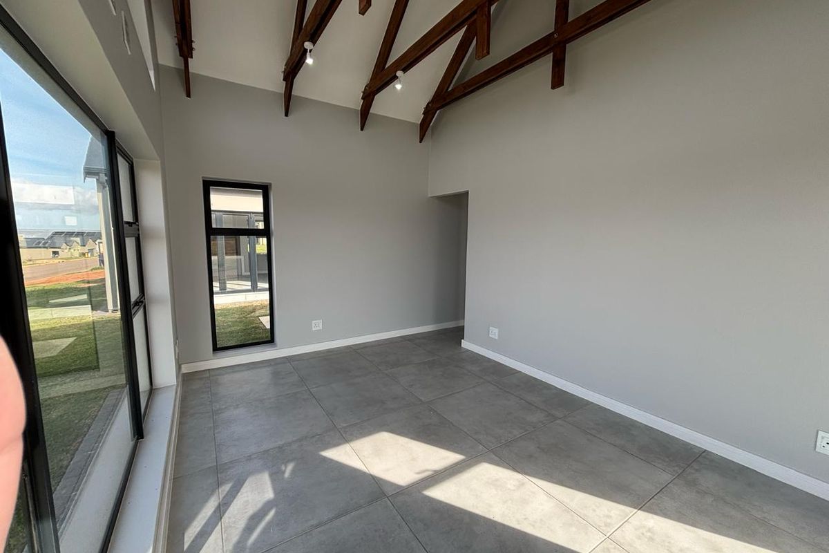 A bright, modern living area featuring high vaulted ceilings with exposed timber trusses, large floor-to-ceiling windows, and neutral tiled flooring.