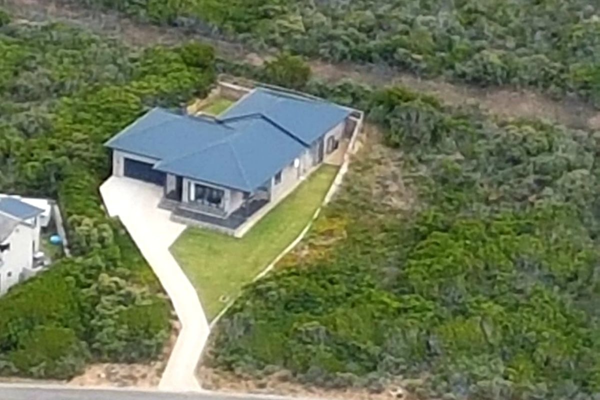 Aerial view of a modern detached house with a dark tiled roof, concrete driveway, and surrounding landscaped garden set within a natural bushland environment.