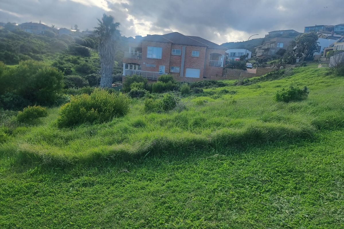 A view of a vacant residential plot with natural grass coverage, looking towards neighboring properties in a hilly coastal or suburban area.