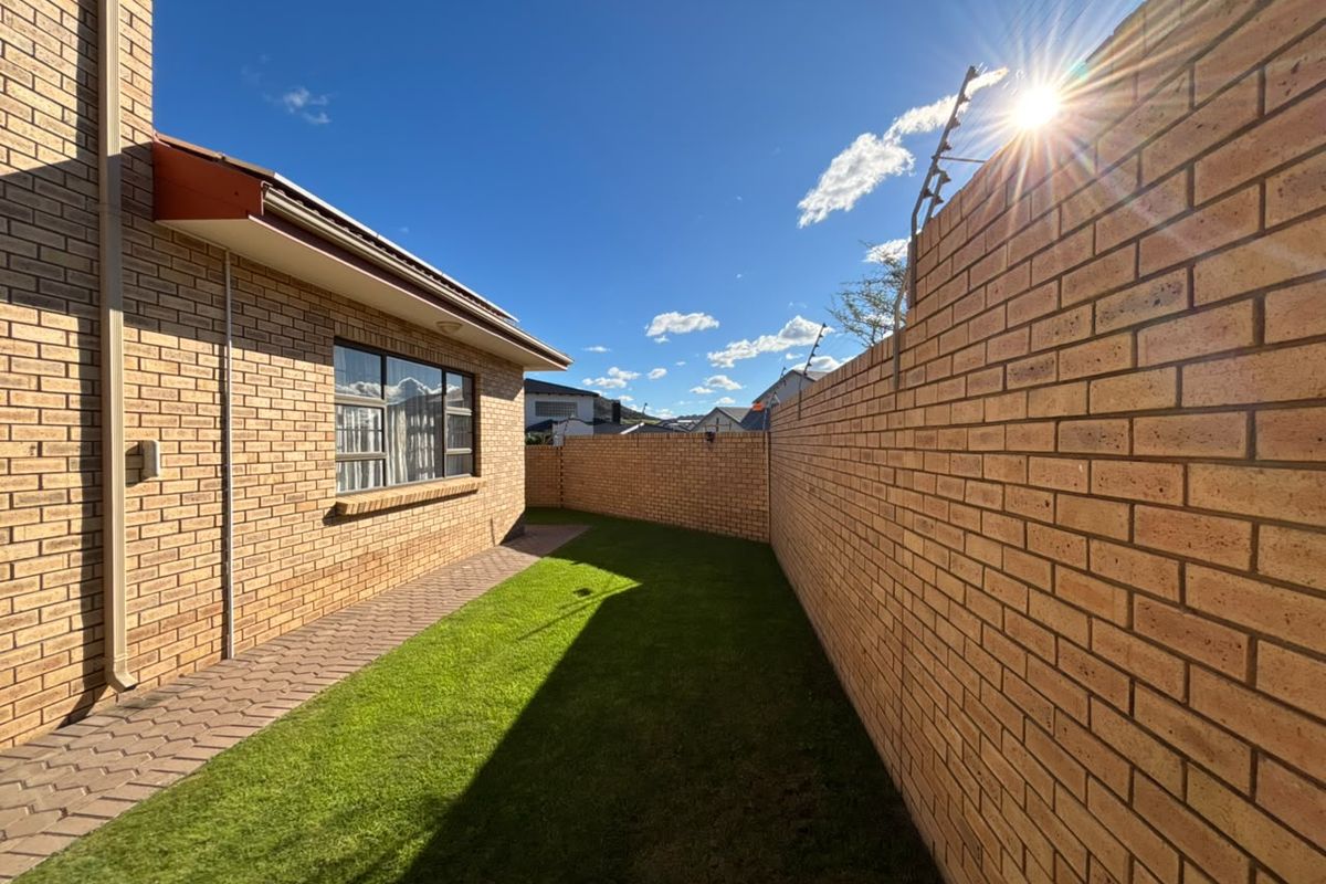 A well-maintained rear garden area featuring a paved walkway, neat lawn, and secure perimeter brick walling with electric fencing.