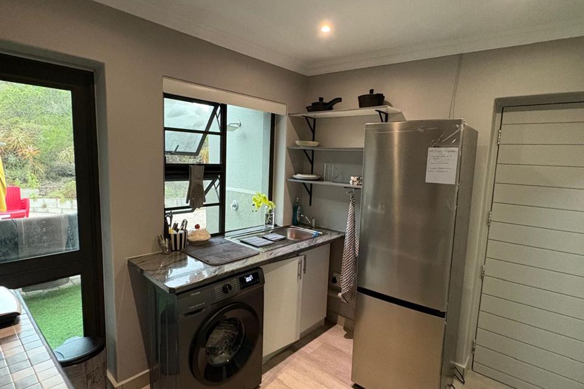 A functional scullery featuring a stainless steel sink, stone countertop, built-in shelving, and space for laundry appliances, with direct access to the outdoor area.