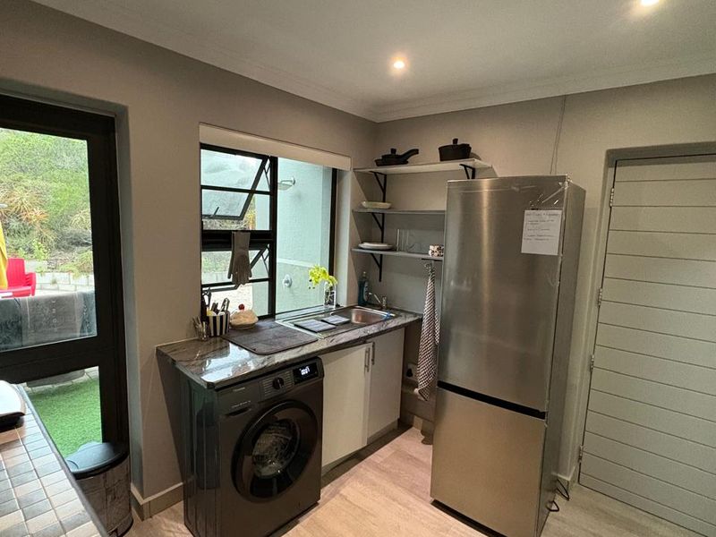 A functional scullery featuring a stainless steel sink, stone countertop, built-in shelving, and space for laundry appliances, with direct access to the outdoor area.