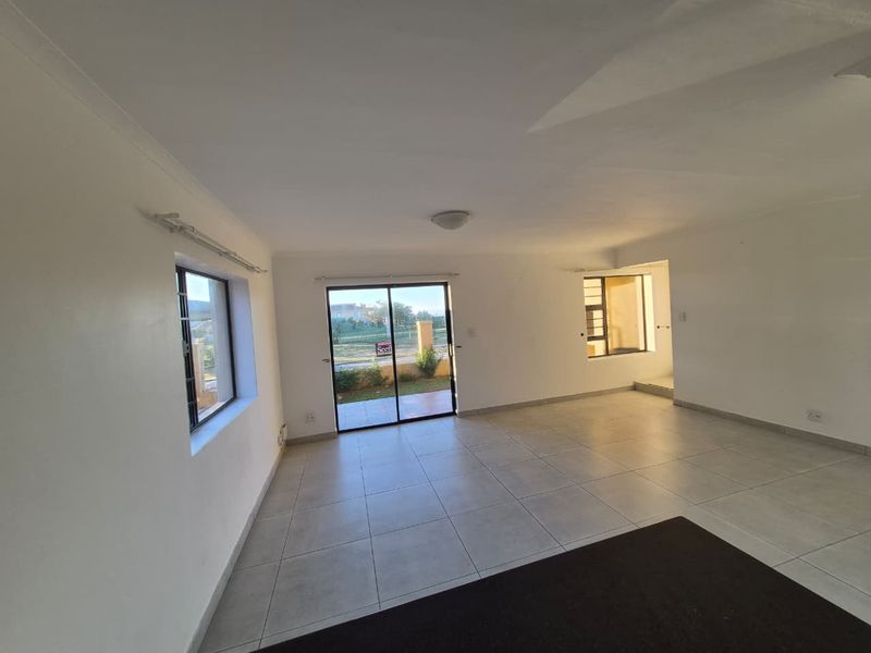 A bright, open-plan living area featuring neutral tiled flooring, large windows with security bars, and a sliding glass door leading to the exterior.