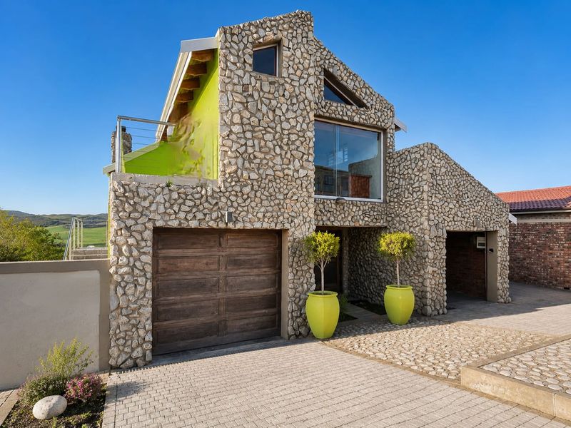 A unique double-story home featuring distinctive stone cladding, a paved driveway, integrated garage, and an upper-level balcony with scenic views.