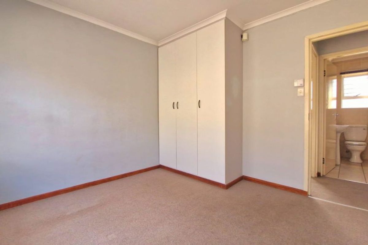 A clean, empty bedroom featuring neutral walls, carpet flooring, and a built-in wardrobe, with an adjoining bathroom visible through the doorway.