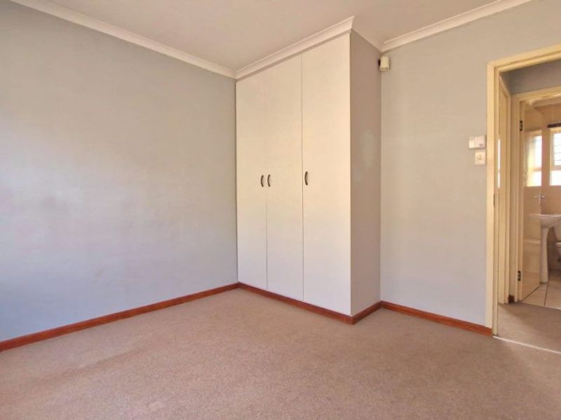 A clean, empty bedroom featuring neutral walls, carpet flooring, and a built-in wardrobe, with an adjoining bathroom visible through the doorway.