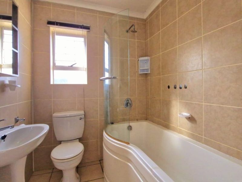 A functional bathroom featuring a pedestal basin, toilet, and a bathtub with an integrated shower and glass screen, finished with neutral wall tiles.