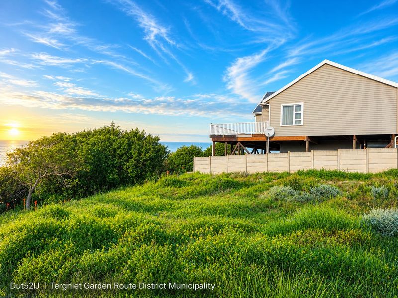 Rear view of the property featuring a raised wooden deck with white balustrades, overlooking a coastal landscape with a precast boundary wall.