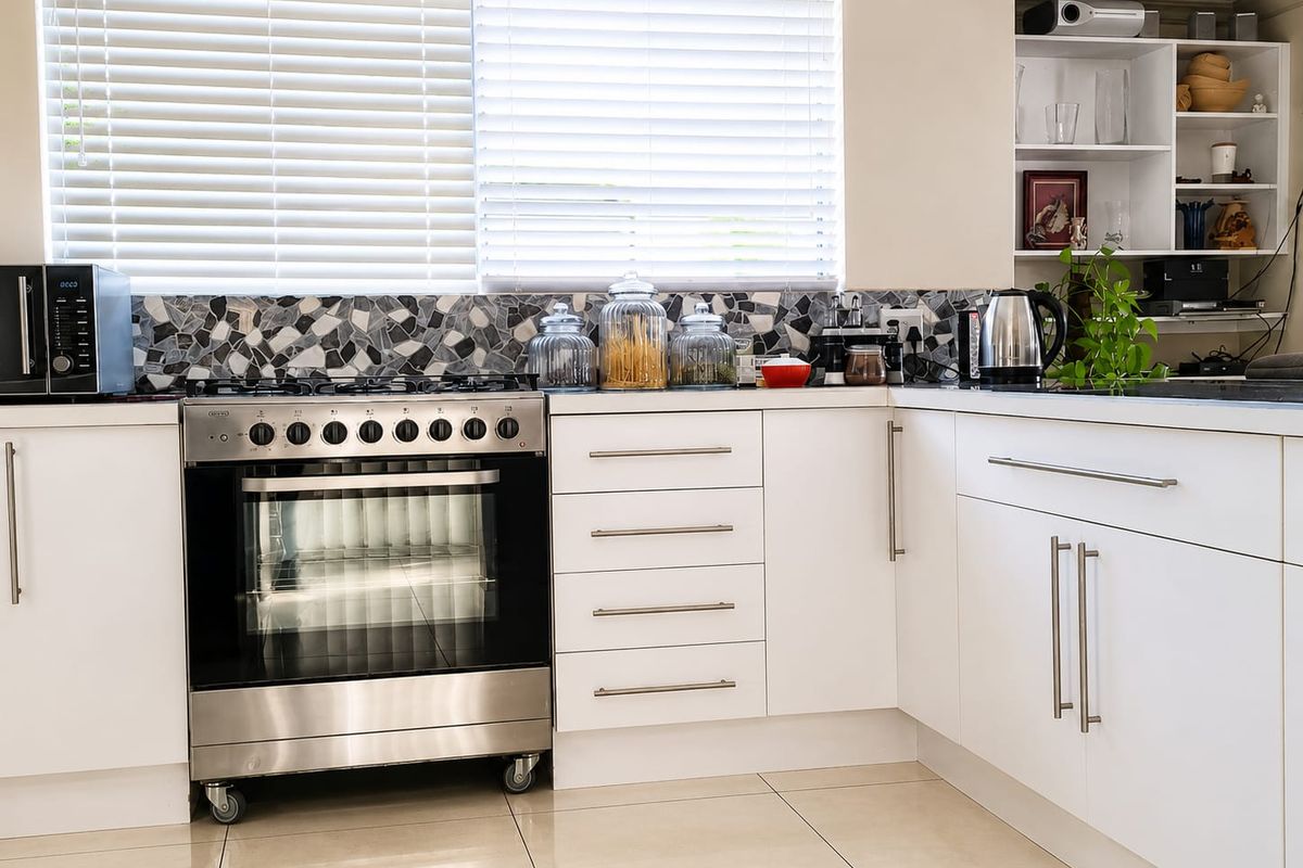 Modern kitchen featuring white cabinetry, stainless steel gas stove with oven, mosaic tile backsplash, and tiled flooring with ample natural light.