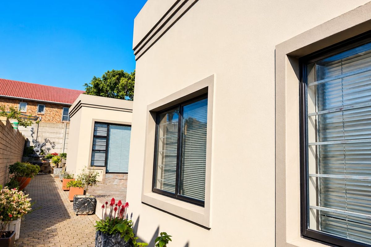 A well-maintained rear exterior view featuring plastered walls, paved pathways, and modern aluminium-framed windows with security bars.