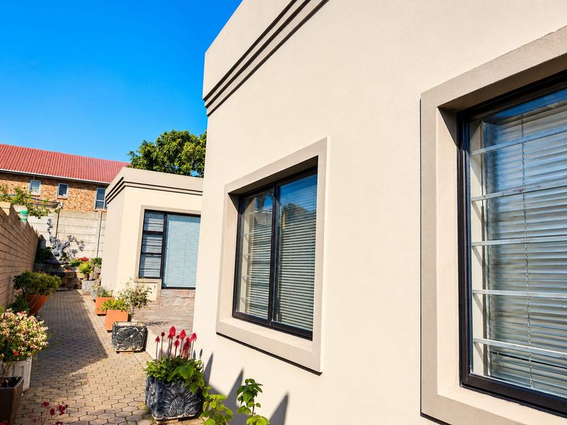 A well-maintained rear exterior view featuring plastered walls, paved pathways, and modern aluminium-framed windows with security bars.