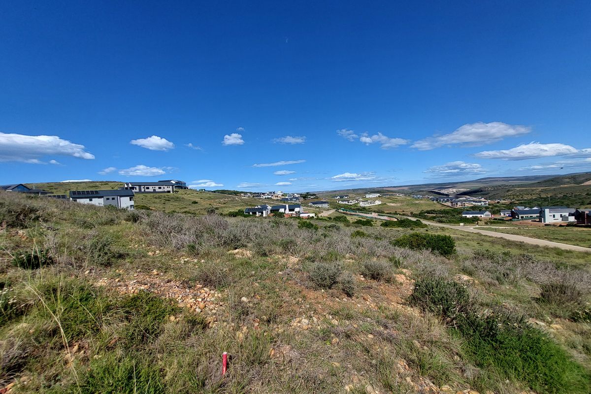 A wide-angle view of a vacant residential plot featuring natural vegetation and a sloped landscape with surrounding neighborhood development in the distance.
