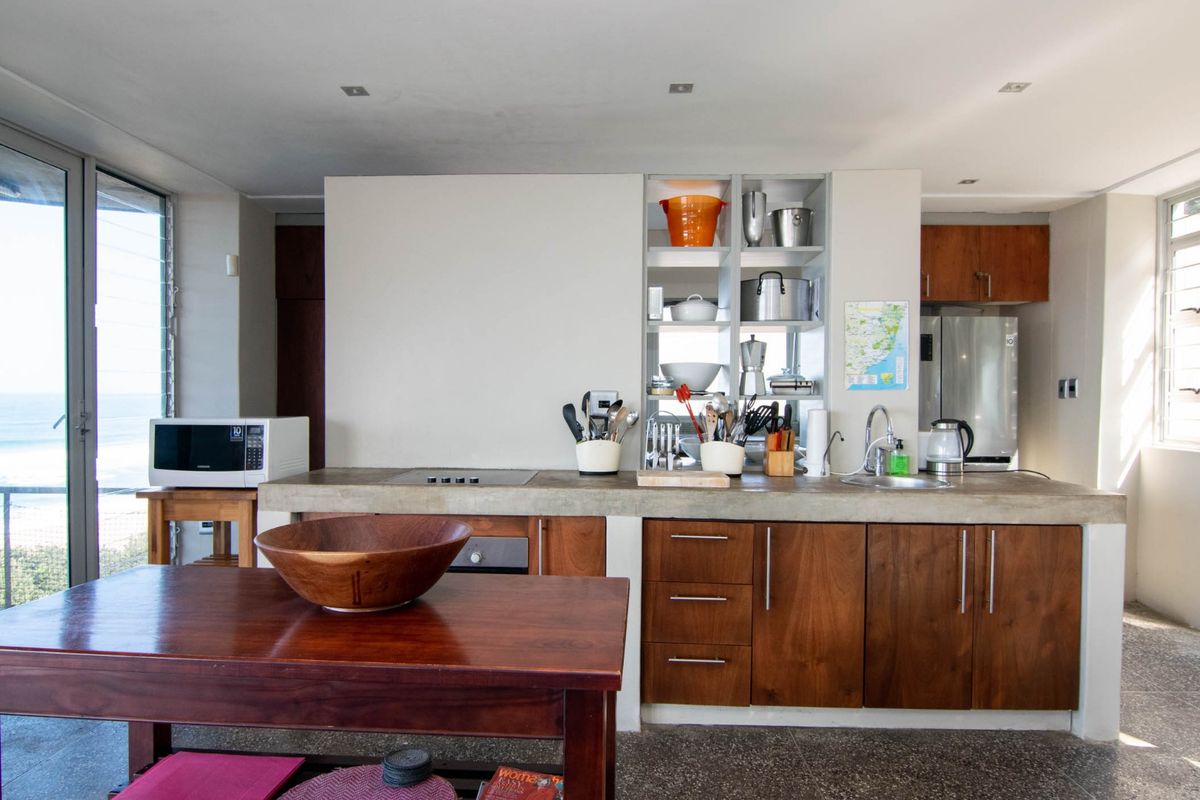 Modern kitchen featuring concrete countertops, wooden cabinetry, built-in cooktop, and integrated shelving, with large windows offering natural light and ocean views.
