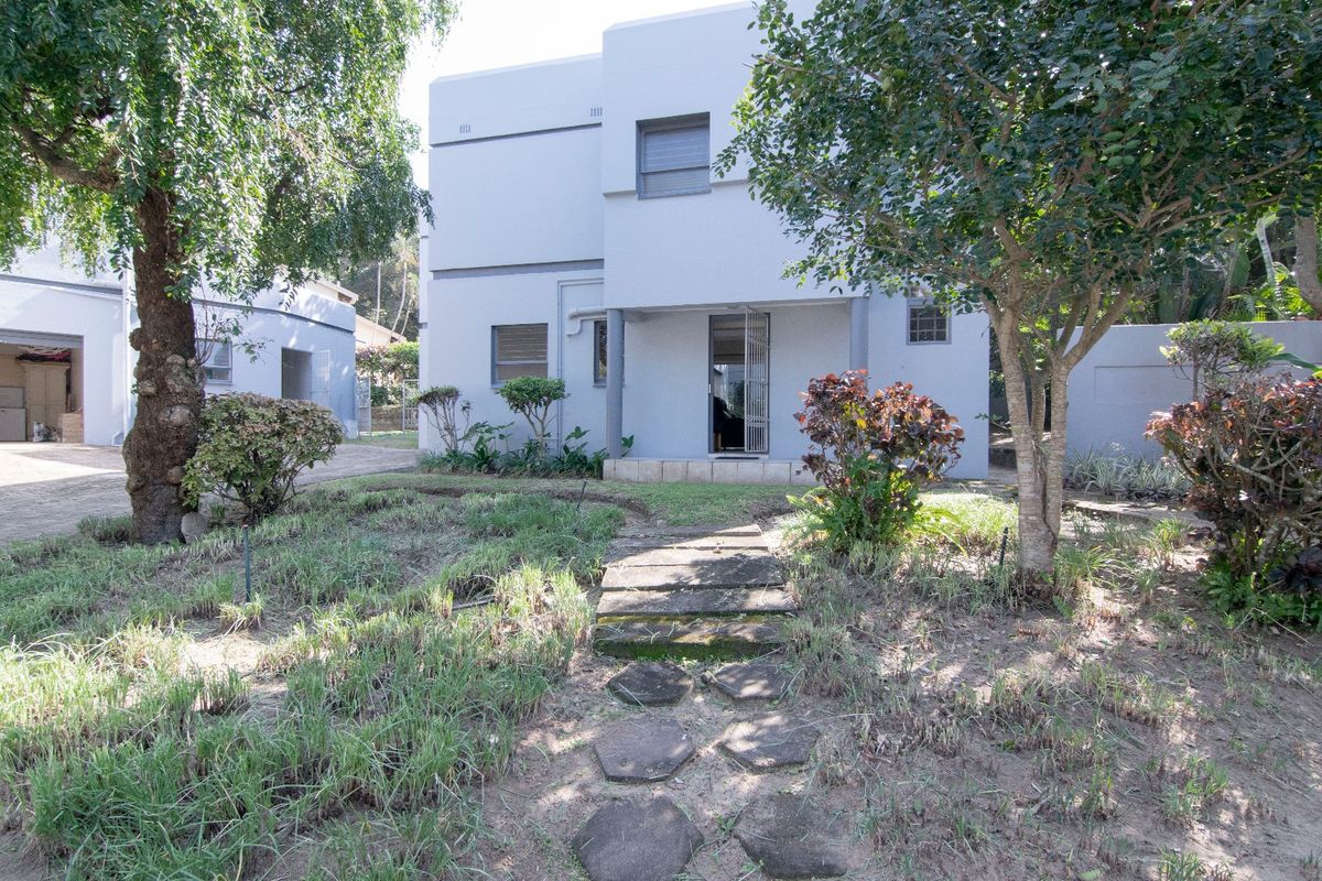 Rear view of a two-story residential property featuring a paved walkway, established trees, and a garden area leading to the building entrance.