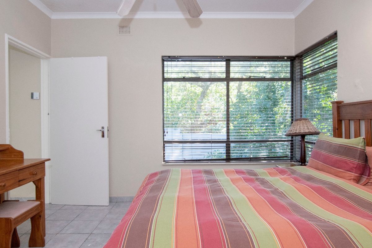 A bedroom featuring tiled flooring, a ceiling fan, and large windows with fitted blinds, offering a view of the garden.