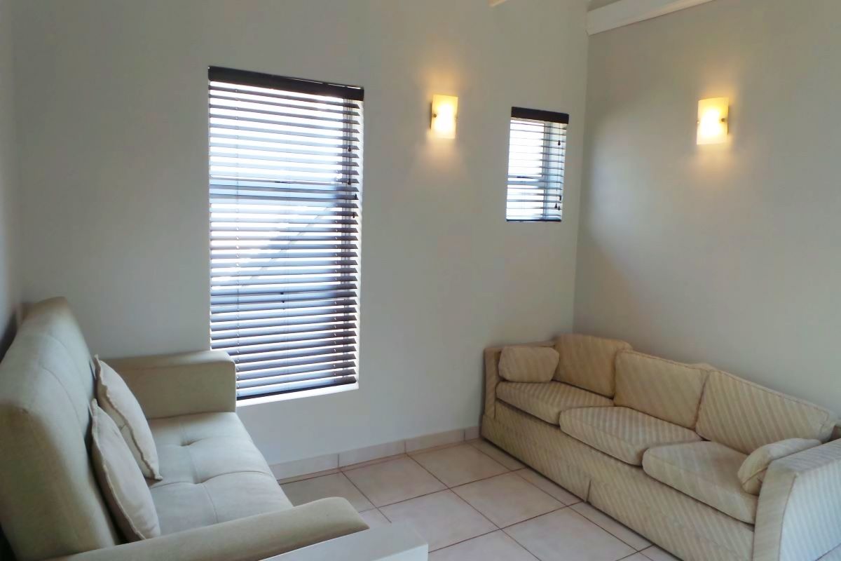 A living room featuring tiled flooring, wall-mounted light fixtures, and windows fitted with horizontal blinds.