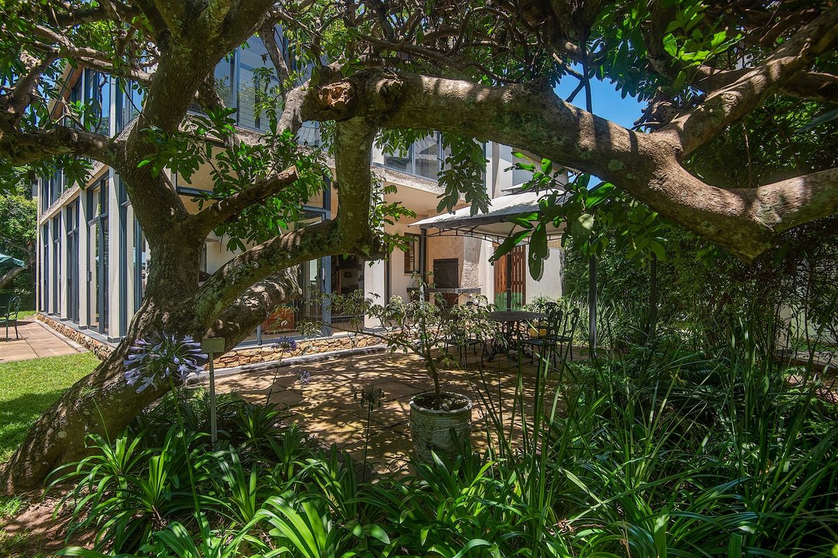 A private rear patio area featuring stone paving, a built-in braai, and large glass sliding doors leading into the home, surrounded by mature garden landscaping.