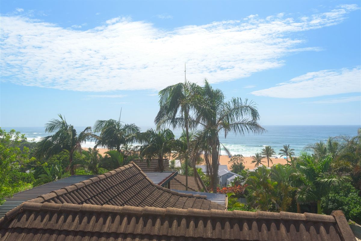 Elevated coastal view overlooking the property roofline, lush tropical vegetation, and the sandy beach with ocean waves.