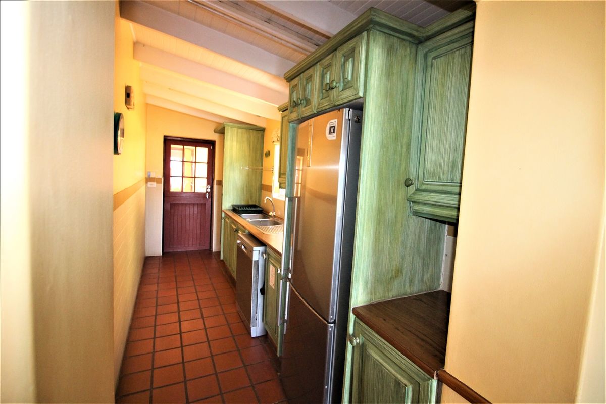 A galley-style kitchen featuring green-toned cabinetry, stainless steel appliances, terracotta floor tiles, and a timber-framed exterior door.
