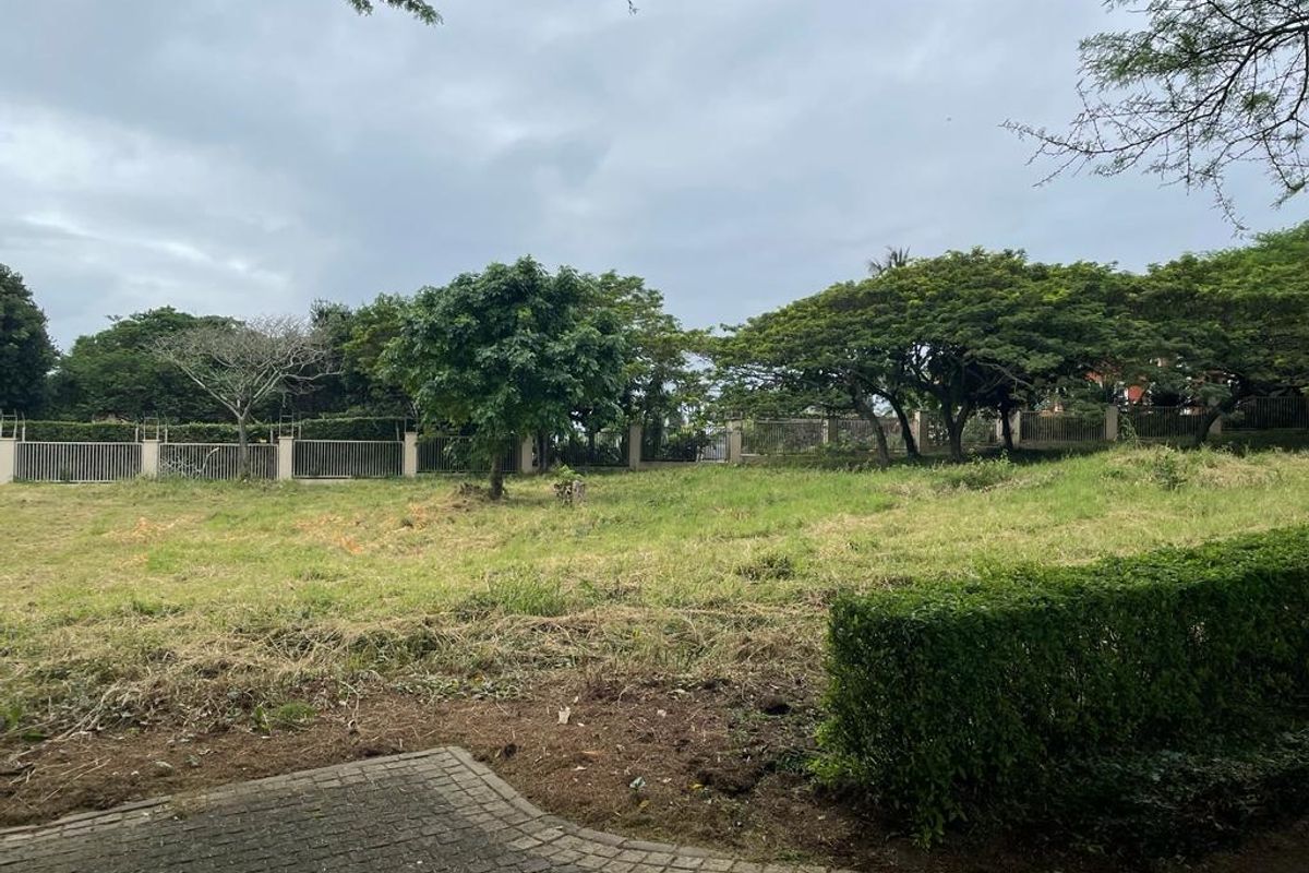 A vacant residential plot featuring established trees, perimeter fencing, and a paved foreground area, offering a clear space for potential development.