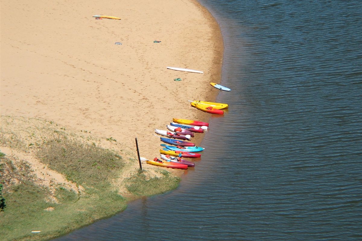 An aerial view of a sandy riverbank featuring a collection of kayaks and water sports equipment along the water's edge.