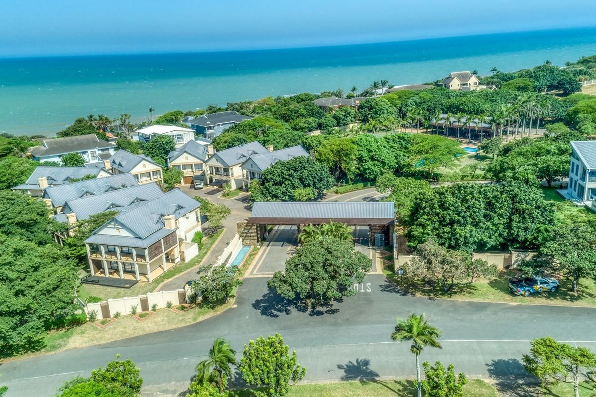 Aerial view of a residential estate entrance featuring a gated access point, paved roads, and surrounding coastal landscape with established trees.