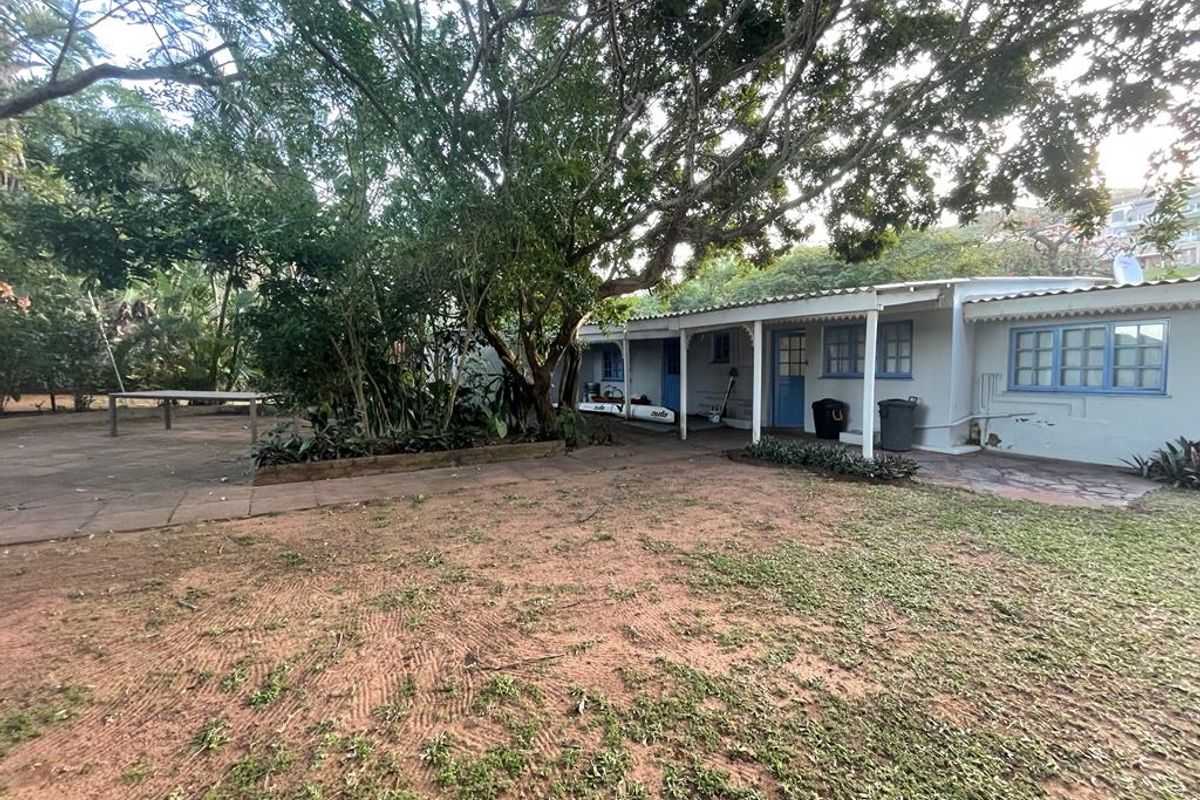 A rear view of the property featuring a covered patio area, established trees, and a garden space with paved sections.