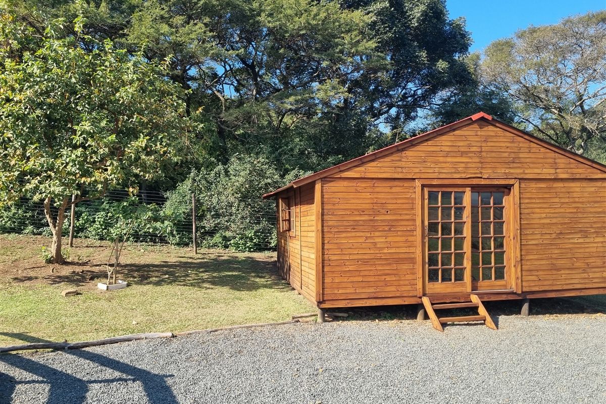 A detached wooden cabin structure featuring French doors and a gravel driveway, set within a lush, tree-lined garden environment.