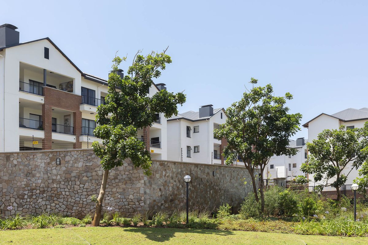 Exterior view of a modern apartment complex featuring multi-story units with balconies, stone boundary walls, and well-maintained landscaped garden areas.