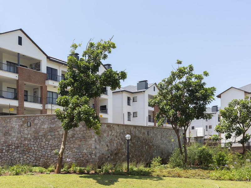 Exterior view of a modern apartment complex featuring multi-story units with balconies, stone boundary walls, and well-maintained landscaped garden areas.