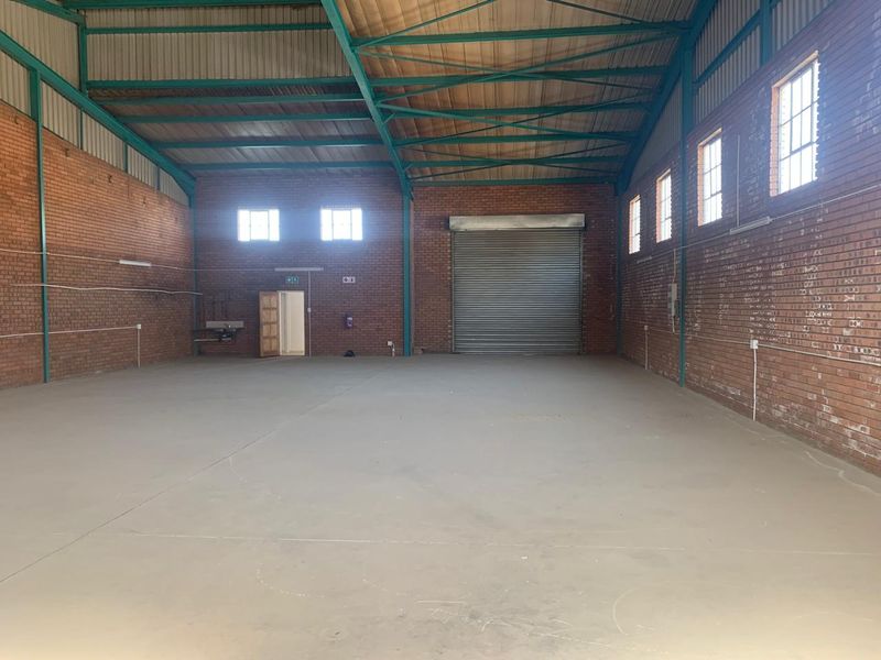 Spacious industrial warehouse unit featuring exposed brick walls, high steel roof trusses, concrete flooring, and a large roller shutter door for easy access.
