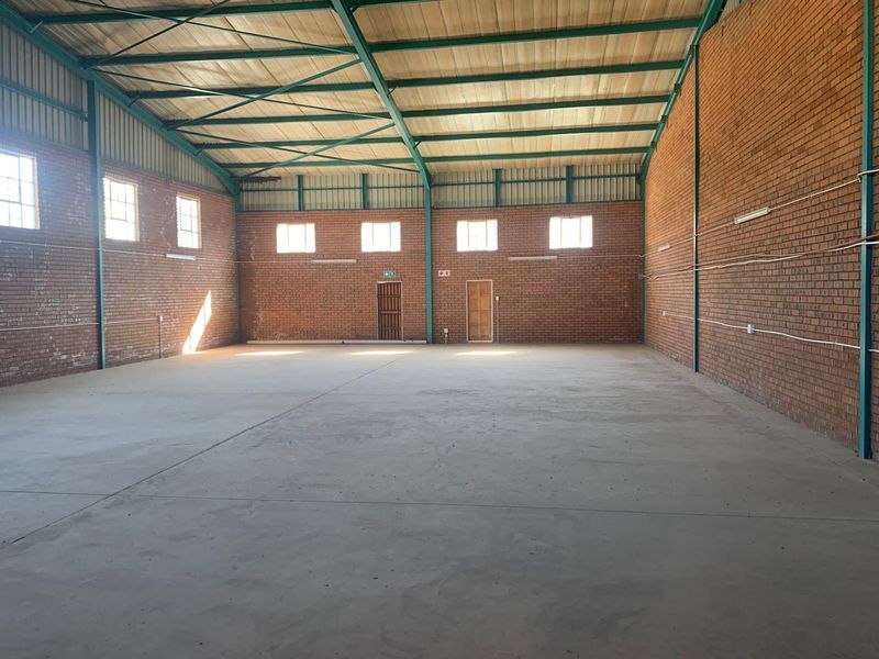 Spacious industrial warehouse unit featuring exposed brick walls, high steel truss ceilings, concrete flooring, and multiple windows providing natural light.