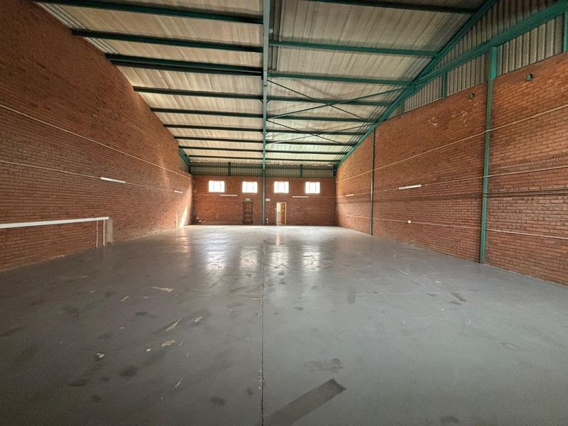 Spacious industrial warehouse unit featuring exposed brick walls, high steel truss ceilings, and durable concrete flooring, suitable for storage or manufacturing operations.