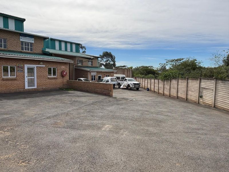 Exterior view of an industrial property featuring a brick facade, metal roofing, and a large asphalt parking area enclosed by a concrete boundary wall.