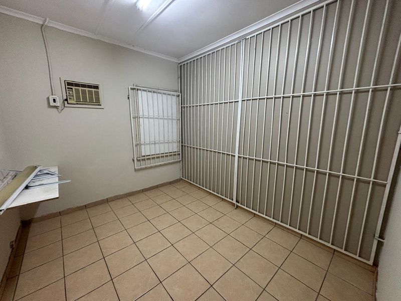 A functional office space featuring tiled flooring, a wall-mounted air conditioning unit, and secure metal security bars covering the window and wall area.